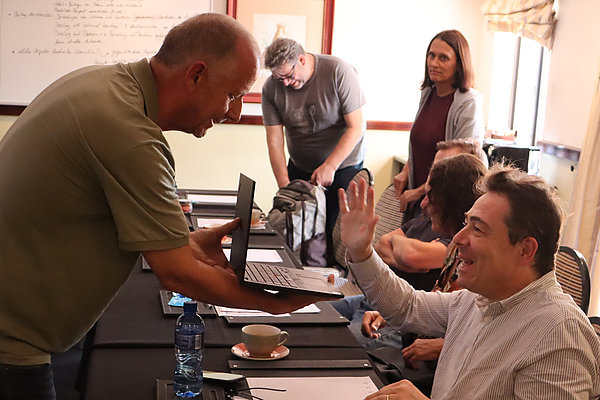 Im Vordergrund hält Stefan Kramer (DVS Brasilien) den Laptop. Frank Kahnert (DVS Tschad) begrüßt den virtuell zugeschalteten Adriano Martins, ein Misereor-Partner aus Brasilien.