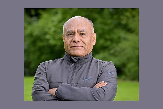 Ein Mann steht mit verschränkten Armen vor einer grünen Wiese. Im Hintergrund sind grüne Bäume verschwommen zu sehen.